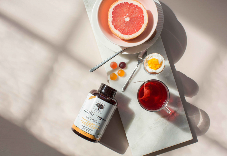 Multi-Vegan Gummies bottle placed on a table with a grapefruit, egg yolk, colourful vitamin gummies, and a red tea drink in natural sunlight.